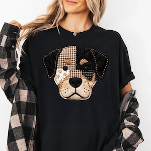 May include: Black t-shirt featuring a patchwork dog face design. The dog's face is made of various fabric patterns in black, brown, and beige, with embroidered details. The shirt has a classic crew neck and short sleeves.