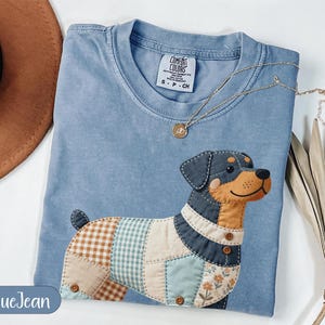 Peut inclure: Un t-shirt bleu jean avec un motif de chien en patchwork. Le chien a un visage noir et beige, et son corps est fait de différents tissus à motifs. Un collier en or repose sur le t-shirt.