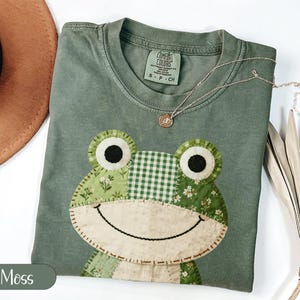 May include: A moss green t-shirt with a frog applique. The frog design features a patchwork of green fabrics, including floral and checkered patterns. A gold necklace with a small pendant rests on the shirt. The shirt is labeled "Comfort Colors".