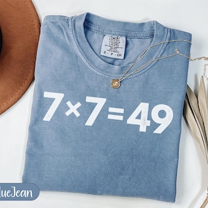 Puede incluir: Una camiseta azul vaquera con la ecuación "7 x 7 = 49" impresa en blanco. La camiseta tiene cuello redondo y una pequeña etiqueta en la parte superior de la espalda. Un collar dorado con un colgante está drapeado sobre la camiseta.
