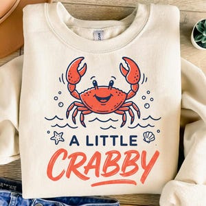 May include: Cream-colored sweatshirt featuring a cartoon crab design in red and navy blue. The text "A LITTLE CRABBY" is written in red script below the crab illustration. A starfish and seashell are also included in the design.