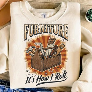 May include: A cream-coloured sweatshirt displaying a graphic of a wooden toolbox filled with carpentry tools. The text "FURNITURE" is above the toolbox, and "It's How I Roll" is below. The design has a vintage, rustic aesthetic.