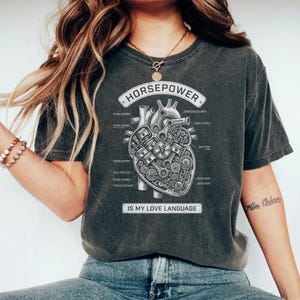 Peut inclure: T-shirt gris foncé avec un diagramme de cœur mécanique et le mot "HORSEPOWER" au-dessus. Le texte "IS MY LOVE LANGUAGE" est en dessous de l'illustration du cœur. Le t-shirt a un style vintage.