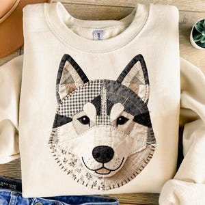 May include: Cream-colored sweatshirt featuring a patchwork design of a husky dog's face. The dog's face is made of various fabric patterns in black, gray, and cream. The sweatshirt has a crew neck and long sleeves. The dog's nose is black, and the eyes are small and dark.
