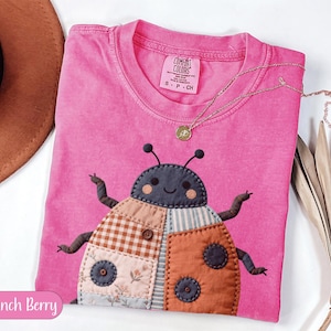 May include: A pink Comfort Colors t-shirt with a patchwork ladybird design. The ladybird has a grey head, brown antennae, and a body made of various fabric patterns. A gold necklace with a small pendant rests on the shirt. The text "Crunch Berry" is at the bottom.