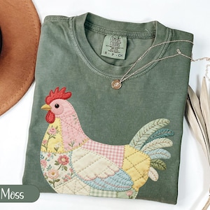 Op de afbeelding: Mosgroen t-shirt met een patchwork kippenontwerp. De kip heeft een rode kam, een roze lichaam en veelkleurige vleugels. Een gouden ketting met een kleine hanger ligt op de shirt. Het shirt is gemaakt van Comfort Colors materiaal.
