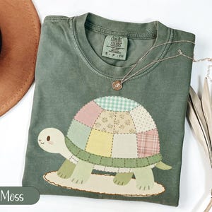 May include: Moss green Comfort Colors t-shirt featuring a patchwork turtle design. The turtle has a light green head and legs, with a shell made of various pastel-colored squares. A gold necklace with a small pendant is draped on the shirt.