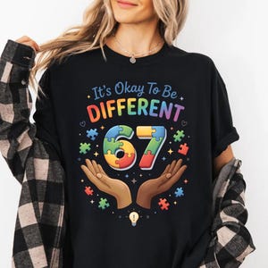 May include: Black t-shirt with the text "It's Okay To Be DIFFERENT" in rainbow colors. The number "67" is designed with puzzle pieces. The design includes hands, puzzle pieces, and a lightbulb.