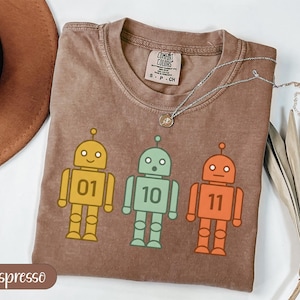 Peut inclure: T-shirt couleur expresso avec trois robots de dessins animés jaunes, verts et oranges. Chaque robot porte un numéro : 01, 10 et 11. Le t-shirt a un col rond et une étiquette Comfort Colors.