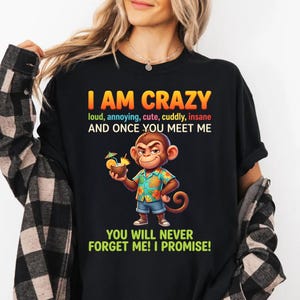 Puede incluir: Camiseta negra con el texto "I AM CRAZY" en naranja y amarillo. La camiseta presenta un mono de dibujos animados sosteniendo una bebida. El texto debajo del mono dice "YOU WILL NEVER FORGET ME! I PROMISE!"