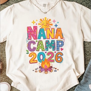 May include: A white short-sleeve t-shirt featuring a colorful graphic with the text NANA CAMP 2026. The design includes a smiling sun, a campfire, and various patterned letters in shades of pink, blue, green, orange, and purple with stitched borders.