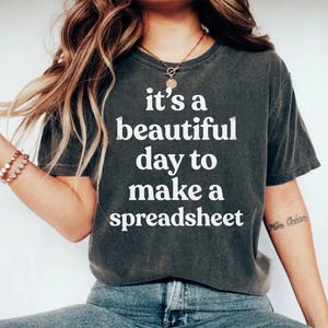 May include: A charcoal grey short-sleeve t-shirt featuring the white text "it's a beautiful day to make a spreadsheet" printed in a bold, sans-serif font across the chest. The garment has a crew neckline and a relaxed, casual fit.