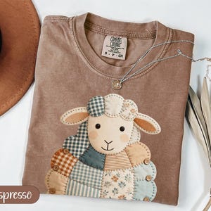 Puede incluir: Camiseta color espresso con un diseño de oveja de patchwork. La oveja tiene una cara color crema, orejas color canela y un cuerpo hecho de varios patrones de tela. Un collar plateado con un pequeño colgante descansa sobre la camisa. La camiseta está etiquetada "Comfort Colors".