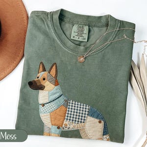 Puede incluir: Una camiseta verde musgo con un diseño de perro patchwork. El perro está hecho de varios patrones de tela en tonos de azul, marrón y blanco. Un collar de oro con un pequeño colgante descansa sobre la camisa. La camiseta tiene cuello redondo y mangas cortas.