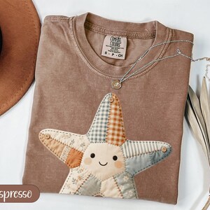 May include: Espresso-colored t-shirt with a patchwork star design. The star features various fabric patterns and button accents. A silver necklace with a pendant rests on the shirt. The shirt is labeled "Comfort Colors" and is size S-CH.