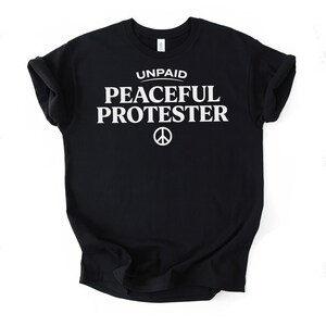 Black t-shirt with white text "UNPAID PEACEFUL PROTESTER" and peace symbol.