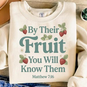 Puede incluir: Sudadera color crema con la frase "By Their Fruit You Will Know Them" en escritura color turquesa. El diseño incluye ilustraciones de fresas y hojas. El texto "Matthew 7:16" está impreso en la parte inferior.
