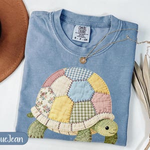May include: A light blue Comfort Colors t-shirt featuring a patchwork turtle design in pastel colors. The turtle has a floral, gingham, and solid fabric shell. A gold necklace with a pendant is draped on the shirt. The text "BlueJean" is on the bottom left.