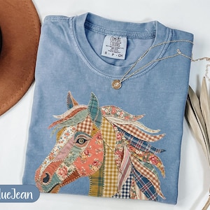 May include: Blue jean colored t-shirt with a patchwork horse head design. The horse's mane and head are made of various floral and plaid fabrics. A gold necklace with a small pendant rests on the shirt. The shirt has a Comfort Colors tag.