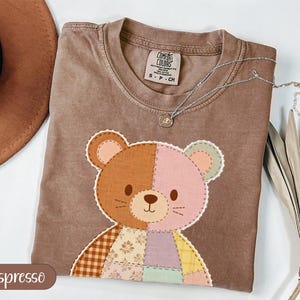 Puede incluir: Camiseta color espresso con un diseño de oso de peluche de patchwork. El oso presenta secciones marrones y rosas, con un patchwork de varios colores. Un collar plateado con un pequeño colgante está drapeado sobre la camiseta. La camiseta tiene cuello redondo y mangas cortas.