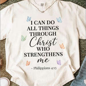 Puede incluir: Camiseta color crema con el texto "I CAN DO ALL THINGS THROUGH Christ WHO STRENGTHENS me - Philippians 4:13." La camiseta presenta gráficos de mariposas en acuarela en azul, verde, naranja y morado.