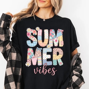 以下が含まれることがあります： パッチワークデザインの「SUMMER VIBES」という文字がプリントされた黒いTシャツ。文字は花柄、ギンガムチェック、ヒョウ柄で装飾されています。デイジーが文字を飾り、テキストは筆記体です。