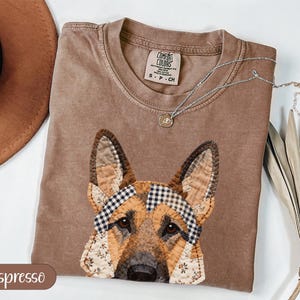 Puede incluir: Camiseta Comfort Colors color Espresso con un diseño de pastor alemán en patchwork. La cabeza del perro está detallada con varios patrones de tela, incluyendo estampados florales y a cuadros. Un collar plateado con un pequeño colgante descansa sobre la camiseta. Talla S-CH.