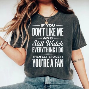 Op de afbeelding: Donkergrijze t-shirt met witte tekst: "IF YOU DON'T LIKE ME AND Still Watch EVERYTHING I DO THEN LET'S FACE IT YOU'RE A FAN". Een casual stijl.