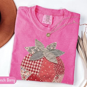 Puede incluir: Una camiseta rosa Comfort Colors con un aplique de fresa. La fresa está hecha de tela de patchwork con patrones rojos, blancos y grises. Un collar de oro con un pequeño colgante descansa sobre la camisa. El texto "Crunch Berry" está en un óvalo rosa.