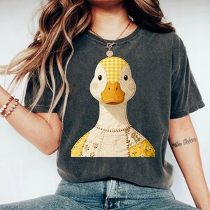 May include: Dark grey t-shirt with a charming duck illustration. The duck has a yellow and white gingham head, a yellow beak, and a patchwork body with floral details. The shirt is a relaxed, casual style.