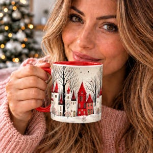 May include: A ceramic mug with a red interior and handle, featuring a winter scene illustration of red and white houses nestled among bare trees against a light background. The design depicts a snowy village landscape with tall, pointed roofs.