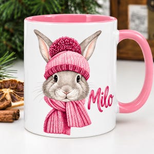 Könnte beinhalten: Ein weißer Keramikbecher mit rosa Innenseite und Henkel, verziert mit einer grafischen Illustration eines grauen Hasen, der eine rosa Strickmütze mit Bommel und einen passenden rosa Schal trägt. Der Name Milo ist in rosa Schreibschrift abgebildet.