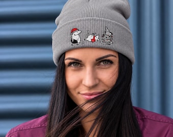 May include: A grey ribbed knit beanie featuring an embroidered graphic of three white mice. One mouse wears a red Santa hat, another wears a red scarf, and the third is tangled in a string of colorful Christmas lights.