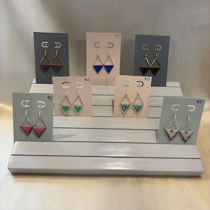 May include: A display of handmade beaded earrings in various colors, including red, blue, green, and gold. Each pair of earrings is a geometric triangle shape and is displayed on a gray or pink card. The earrings are on a white tiered display.