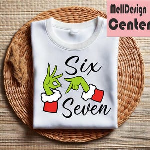 Pu&ograve; includere: Felpa bianca con le parole "Six Seven" in corsivo nero. Il design presenta mani verdi con polsini rossi e bianchi, che imitano un popolare personaggio delle vacanze. La felpa &egrave; piegata su una superficie intrecciata marrone.