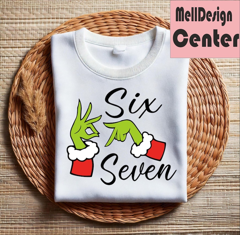 Pu&ograve; includere: Felpa bianca con le parole "Six Seven" in corsivo nero. Il design presenta mani verdi con polsini rossi e bianchi, che imitano un popolare personaggio delle vacanze. La felpa &egrave; piegata su una superficie intrecciata marrone.