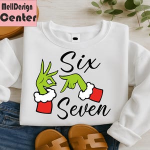 Pu&ograve; includere: Felpa bianca con le parole "Six Seven" in corsivo nero. Il design include mani verdi con polsini rossi e bianchi, che formano i numeri sei e sette. La felpa &egrave; esposta su uno sfondo di jeans.