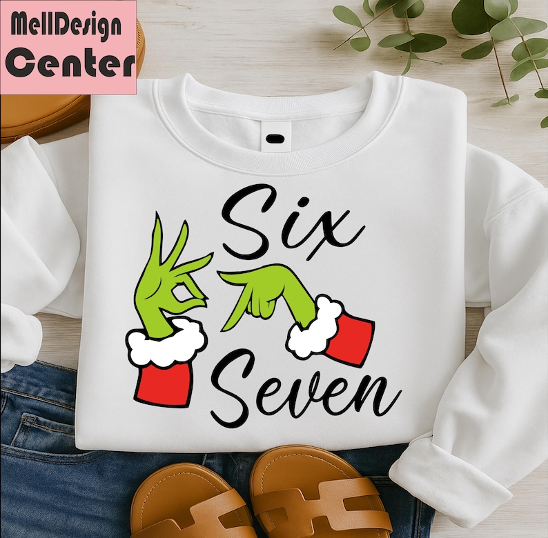 Pu&ograve; includere: Felpa bianca con le parole "Six Seven" in corsivo nero. Il design include mani verdi con polsini rossi e bianchi, che formano i numeri sei e sette. La felpa &egrave; esposta su uno sfondo di jeans.