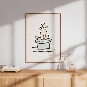 Pode incluir: Uma impressão artística emoldurada apresenta uma ilustração minimalista de um gato sentado em um chapéu. A obra de arte é em um estilo de linha simples com um gato bege claro e um chapéu verde claro. A moldura é marrom quente.
