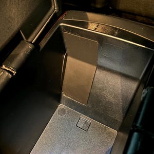 Könnte beinhalten: Geöffnete schwarze Autokonsole mit rechteckiger Metallplatte und quadratischer Vertiefung. Das Innere ist dunkel gehalten. Leichte Gebrauchsspuren sind erkennbar.