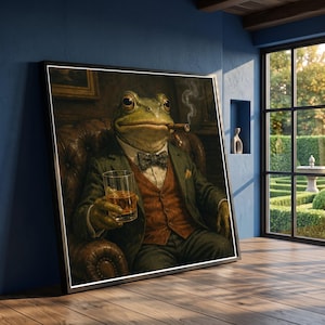 May include: A framed wall art print depicting a green frog dressed in a dark green suit jacket, a brown vest, and a patterned bow tie. The frog is seated in a leather armchair, holding a glass of amber liquid and smoking a cigar with visible smoke.