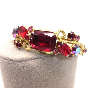 Puede incluir: Un brazalete dorado con una gran gema rectangular de color rojo rubí en el centro. El brazalete está adornado con gemas rojas y moradas más pequeñas, algunas con efectos iridiscentes. El diseño tiene un patrón floral o de vid.