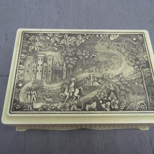 May include: Rectangular cream-colored trinket box with a detailed black and white relief scene. The lid features a castle, figures on horseback, and rural buildings. The box has a decorative, textured border.