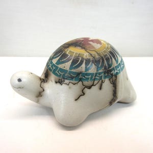 Może przedstawiać: Ceramiczna figurka żółwia o gładkim, kremowobiałym korpusie z ciemnymi, cienkimi, rozgałęzionymi wzorami spękań. Skorupa ozdobiona jest kolistym pasem turkusowych geometrycznych kształtów, żółtym pierścieniem oraz czerwono-białym wzorem pośrodku.