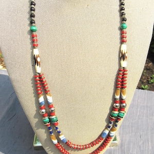 Puede incluir: Un collar de varias capas con cuentas negras, rojas, blancas, azules y verdes. El collar presenta cuentas cilíndricas color hueso con patrones marrones y detalles plateados. El collar se exhibe en un maniquí.