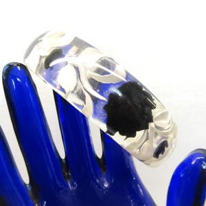 May include: A clear resin bangle bracelet featuring an embedded design of white leaves and black floral shapes. The circular accessory has a smooth, polished surface and a thick, rounded profile, showcasing a botanical pattern throughout.