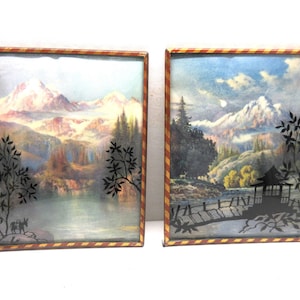 May include: Two framed landscape art pieces. Each features a mountain scene with a lake, trees, and a decorative border. One has a silhouette of a bridge and a structure, the other has a silhouette of people and animals.
