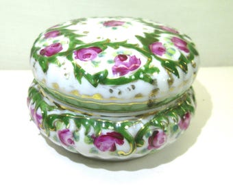 May include: A round, white porcelain trinket box with a lid, decorated with pink roses and green foliage. The floral pattern is set against a white base, with gold detailing. The box has a vintage aesthetic.