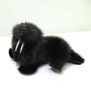 May include: A small, black, plush walrus figurine with two prominent white tusks. The toy has a furry texture and is posed lying down. The figurine is on a white surface.