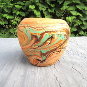 Op de afbeelding: Een afgeronde keramische pot met een gemarmerd patroon in wervelende kleuren van terracotta oranje, mintgroen, donkerbruin en zwart. Het oppervlak heeft een gladde afwerking met een brede opening aan de bovenkant en een naar de basis toelopend lichaam.